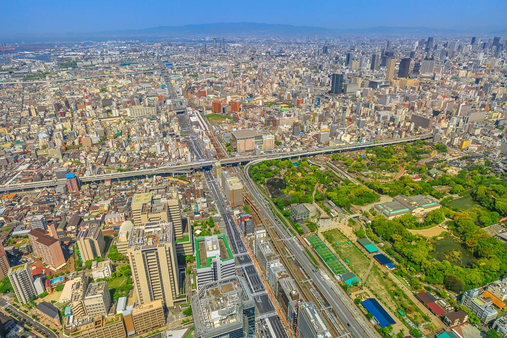 Panorama de Osaka visto do alto do Abeno Harukas, com muitos prédios e uma grande área verde ao lado de vias elevadas.