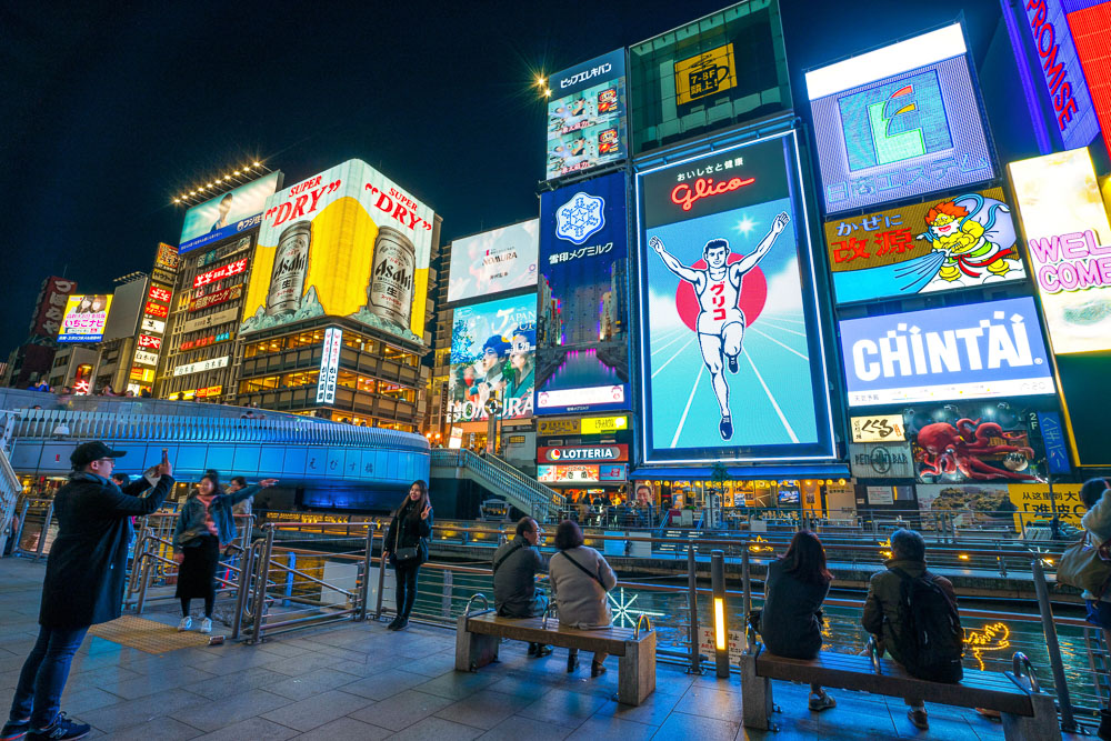 : Letreiros luminosos de Dotonbori à noite, com o famoso Glico Man e pessoas na beira do canal, Osaka. Legenda: O Glico Man em
