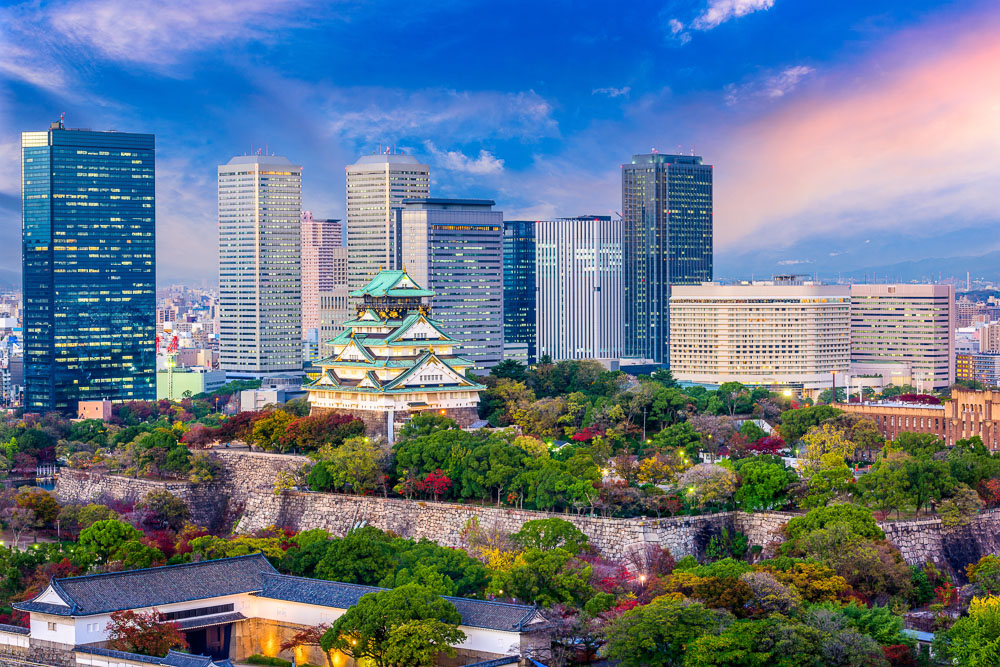 : Castelo de Osaka cercado por área verde e prédios modernos ao fundo, visto de longe no fim do dia.