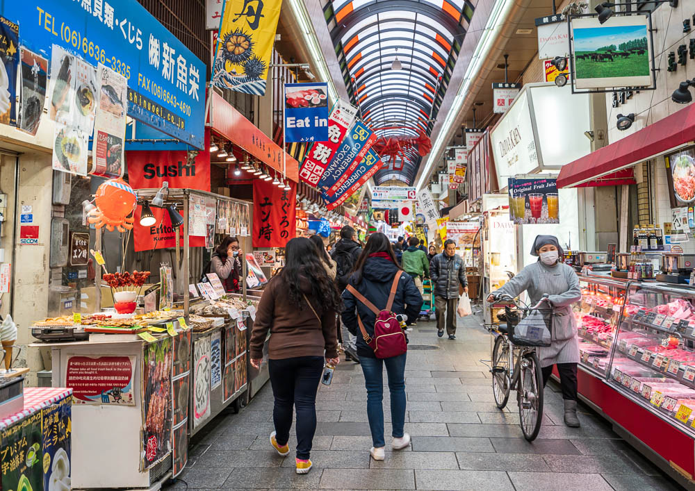 Corredor do Kuromon Market em Osaka, com barracas de comida, letreiros coloridos e pessoas caminhando.