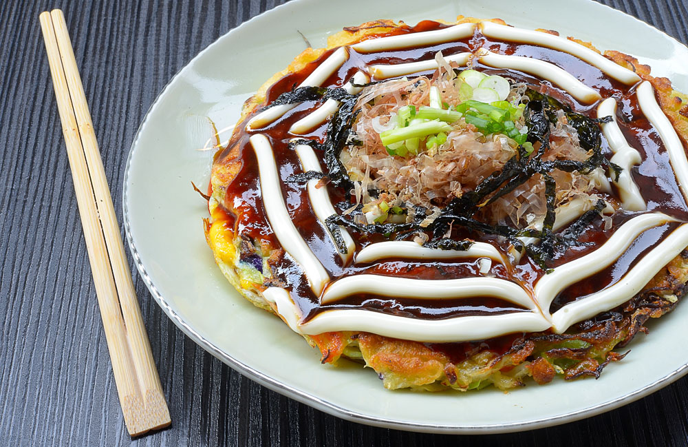 Okonomiyaki servido em prato branco, coberto com molho escuro, maionese e acompanhamentos.
