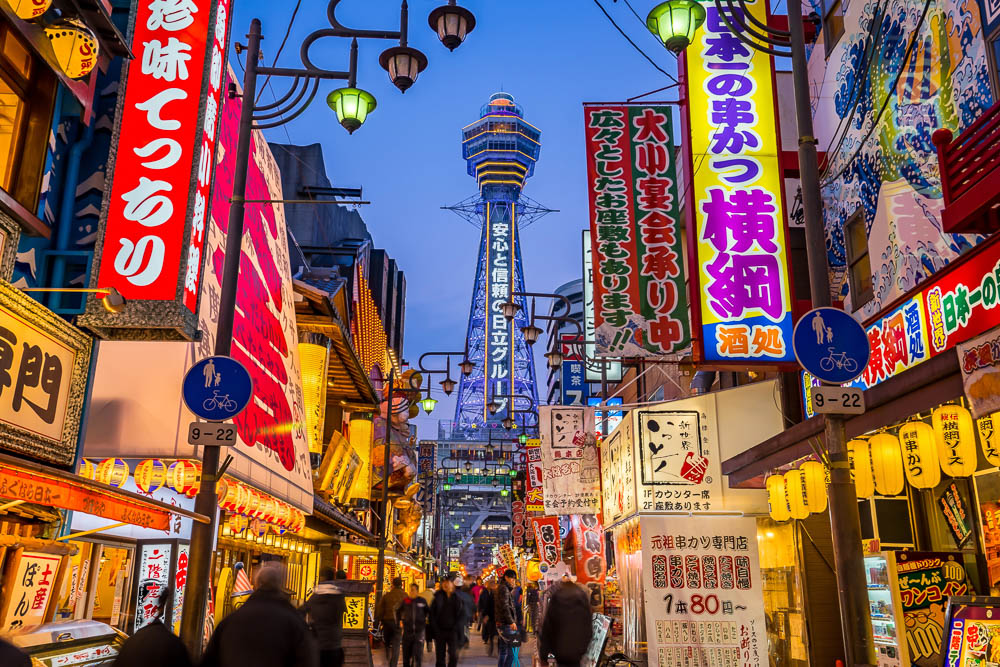 Rua iluminada de Shinsekai em Osaka, com letreiros coloridos e a Torre Tsutenkaku ao fundo.