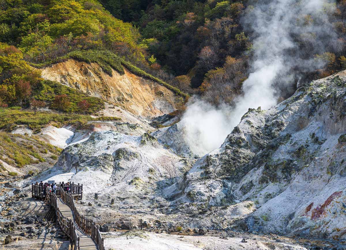 Jigokudani, o Vale do Inferno, com trilha de madeira, vapor geotérmico e encostas claras.