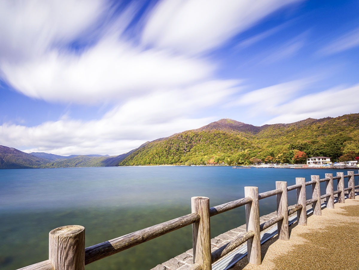 Lago Shikotsu próximo a Sapporo, com águas calmas, montanhas verdes e cerca de madeira à beira do caminho.