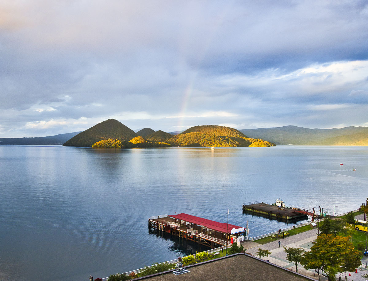Lago Toya visto do alto, em Hokkaido, com ilhas ao centro e píer à beira da água.