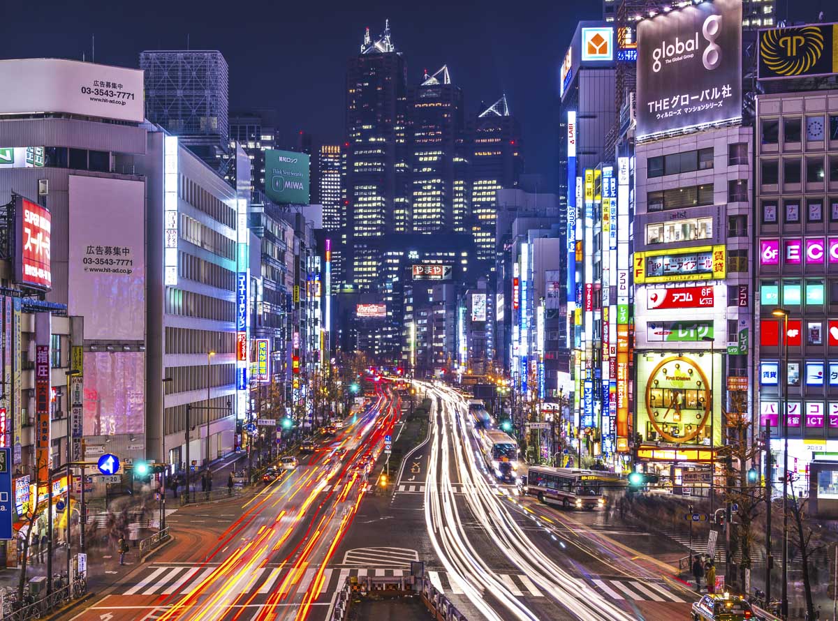 Avenida iluminada em Tokyo à noite, com trilhas de luz dos carros e prédios cheios de letreiros.