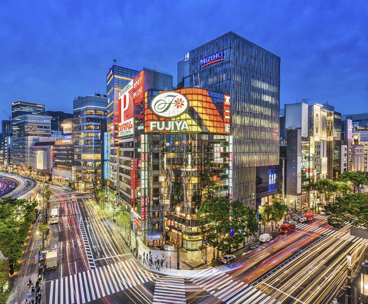 Cruzamento iluminado de Ginza à noite, em Tokyo, com prédios modernos e tráfego intenso.