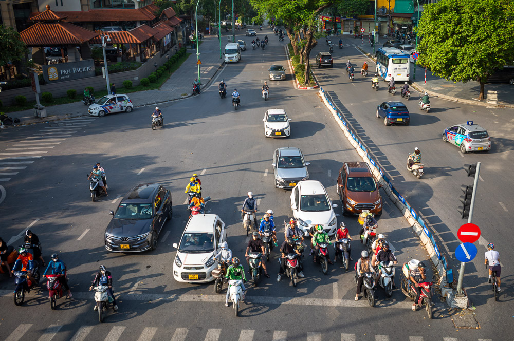 Cruzamento movimentado no Vietnã, com muitas motos, carros e ônibus atravessando a avenida.