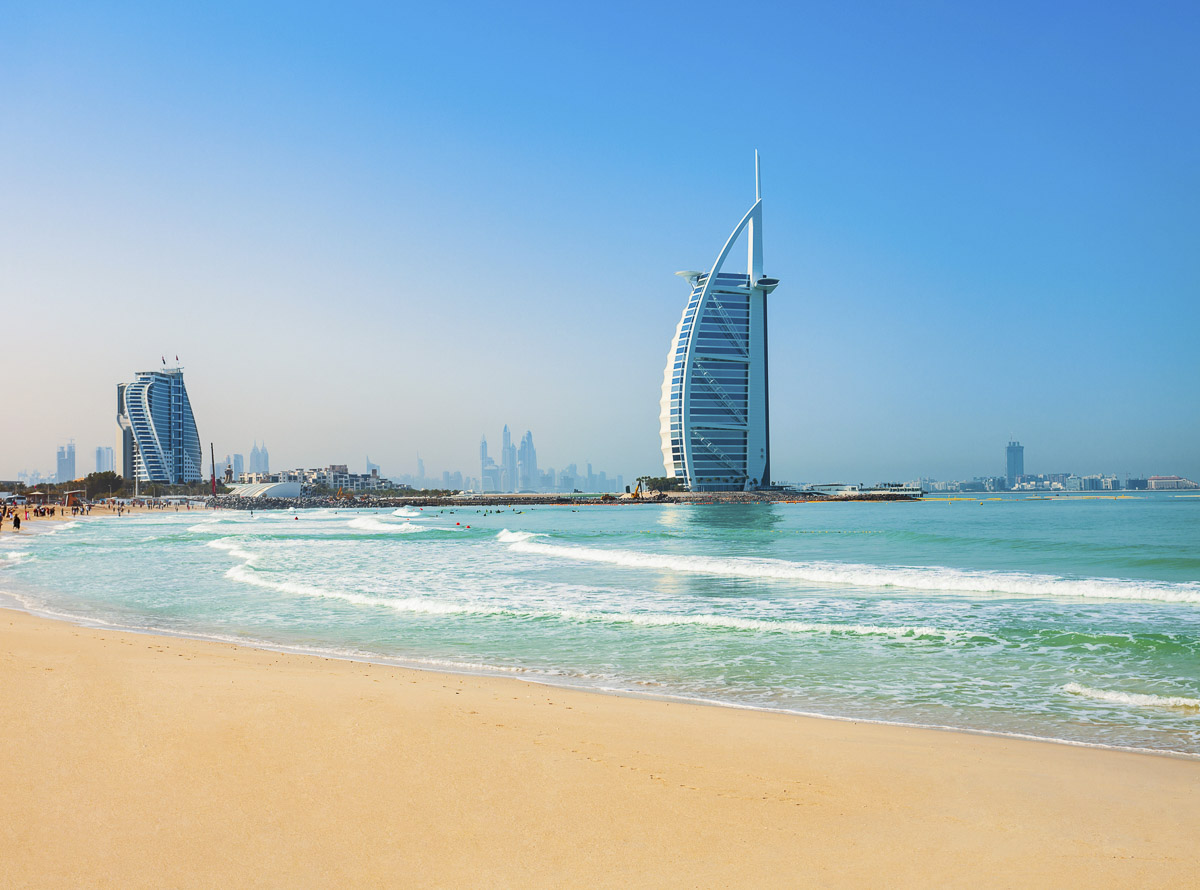 Faixa de areia da Jumeirah Public Beach com o Burj Al Arab ao fundo e mar azul claro.