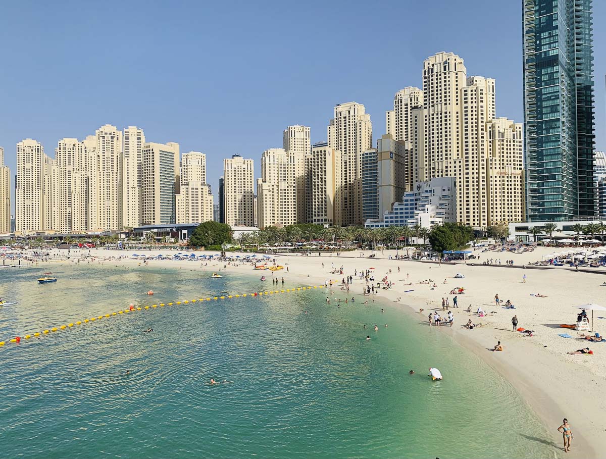 Faixa de areia e mar verde-azulado em The Beach, em Dubai, com banhistas e prédios altos ao fundo.