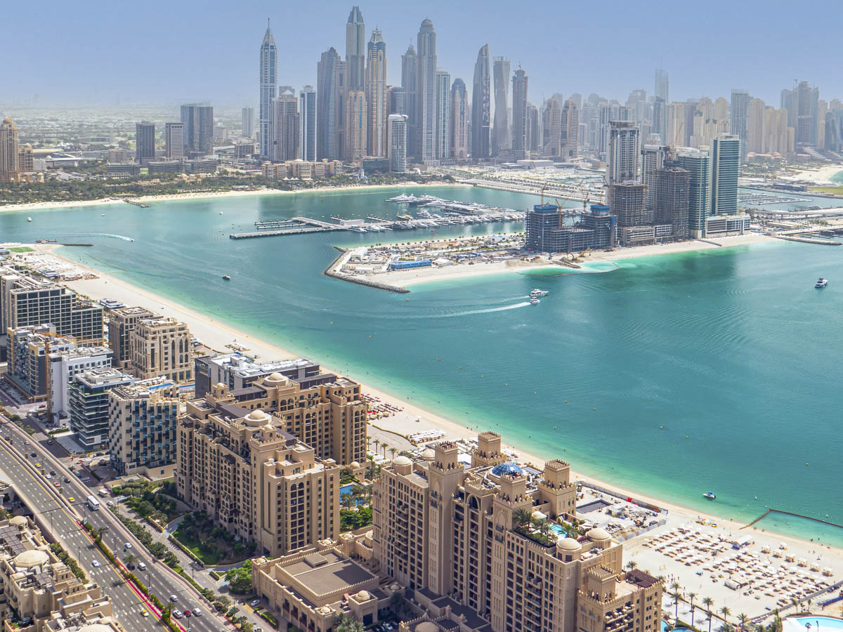 Vista aérea do mar azul e da orla com prédios altos, mostrando as praias de Dubai e a marina ao fundo.