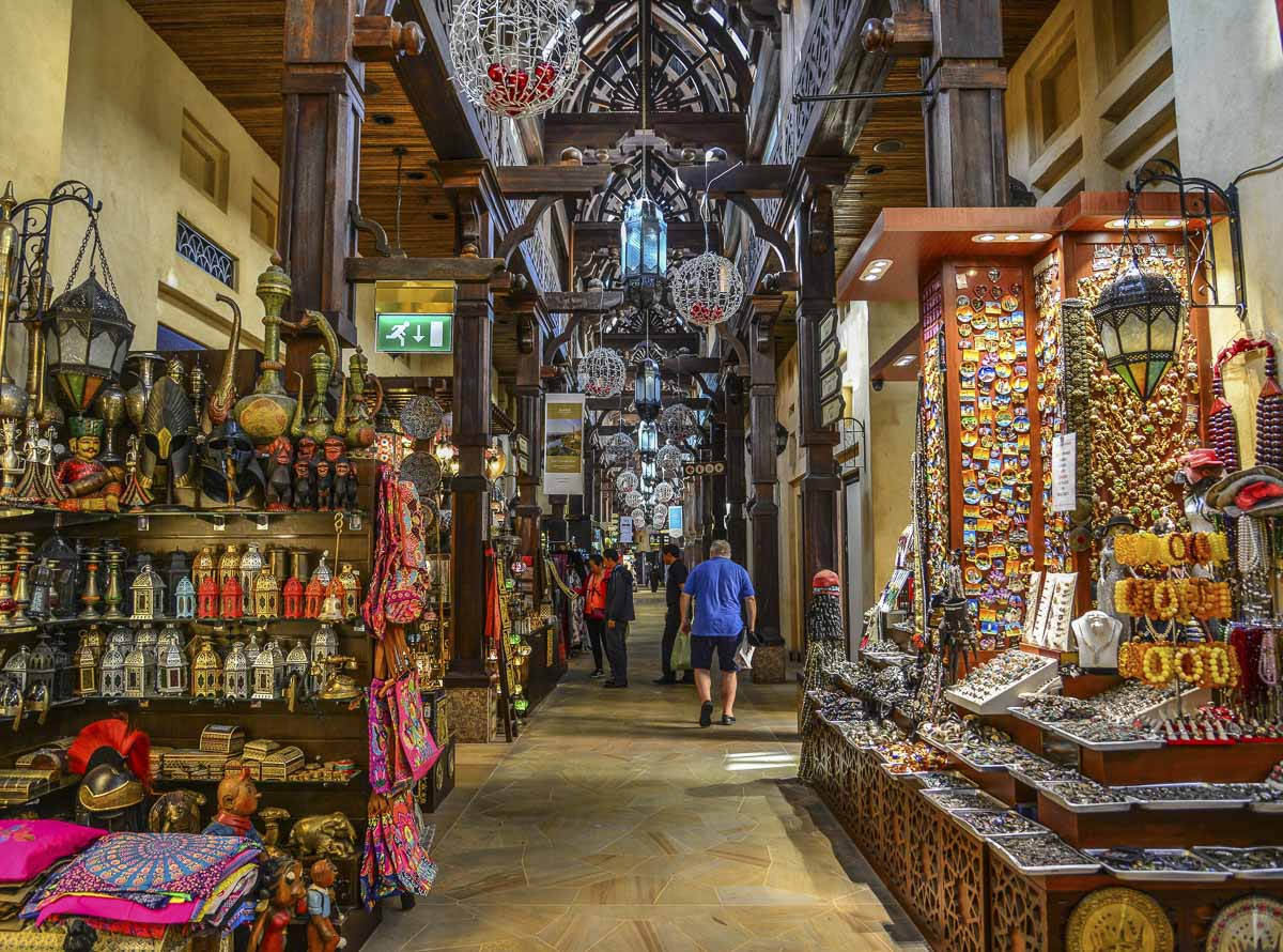 Corredor do Souk Madinat Jumeirah com lojas de artesanato, lanternas e visitantes caminhando.