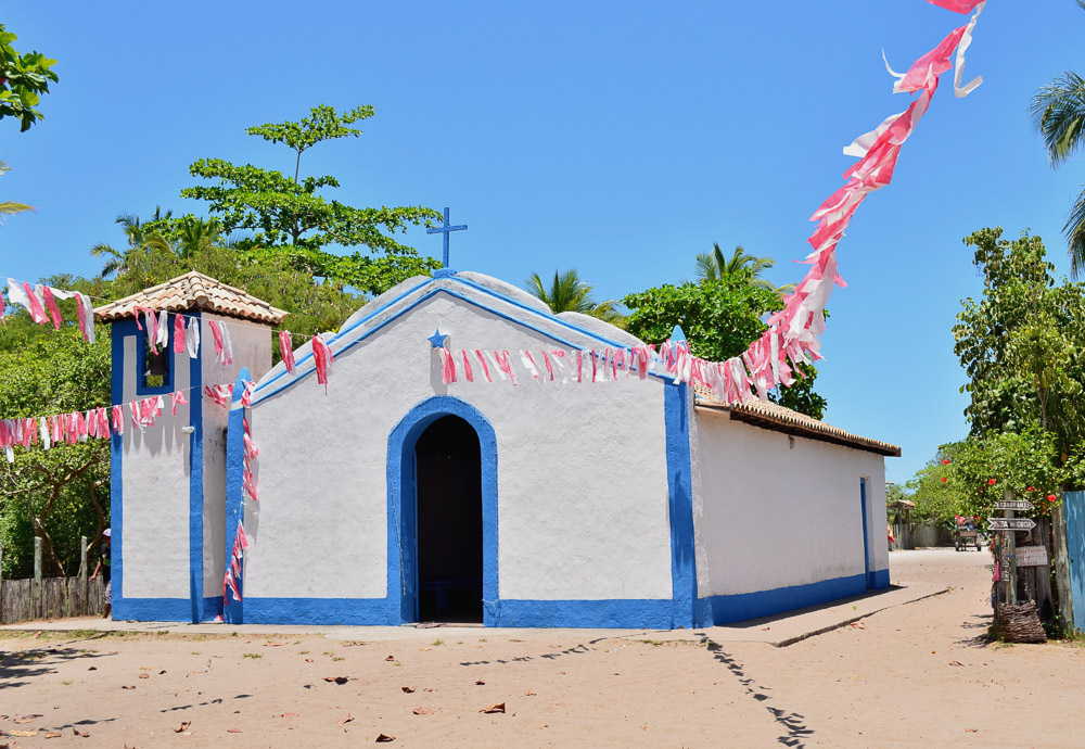 Igreja de São Sebastião, branca e azul em rua de areia, com bandeirinhas coloridas e céu bem azul em Caraíva.