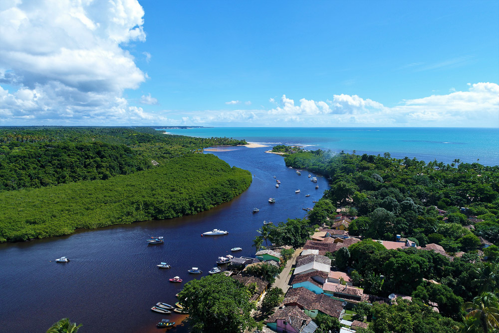 Vista aérea do Rio Caraíva com barquinhos, manguezal e o vilarejo ao lado do mar.