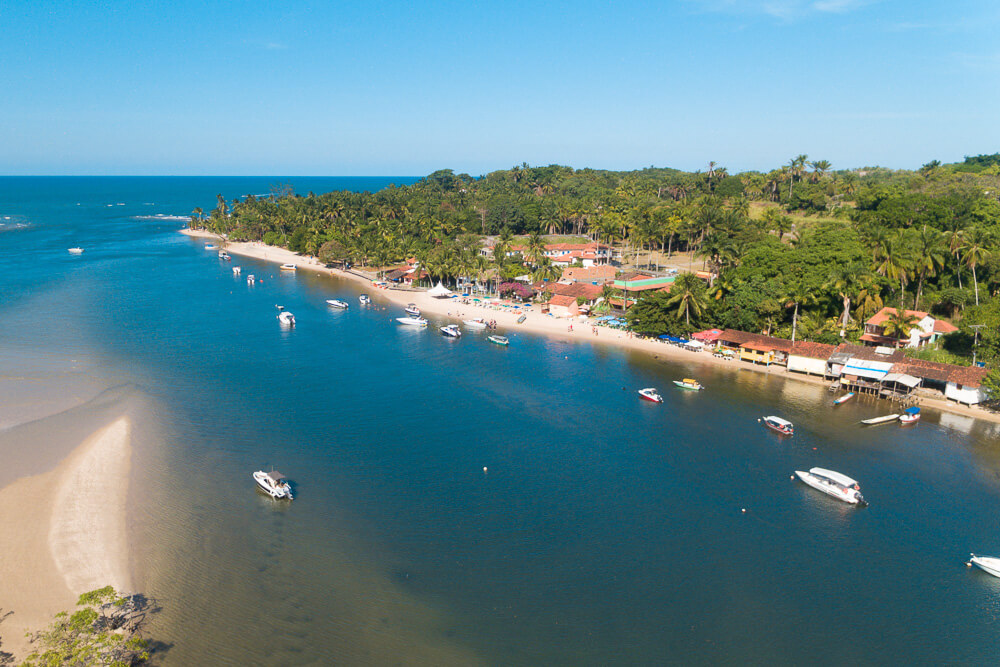 Praia Boca da Barra em Boipeba vista do alto, com rio calmo, barcos e vilarejo ao fundo.