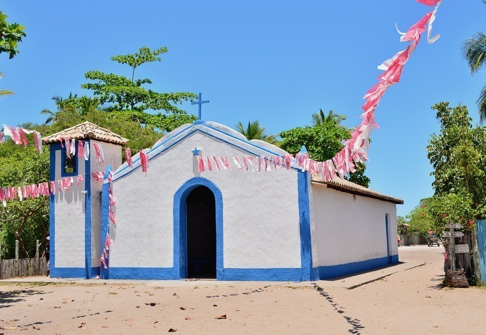 Igreja de São Sebastião, branca e azul em rua de areia, com bandeirinhas coloridas e céu bem azul em Caraíva.