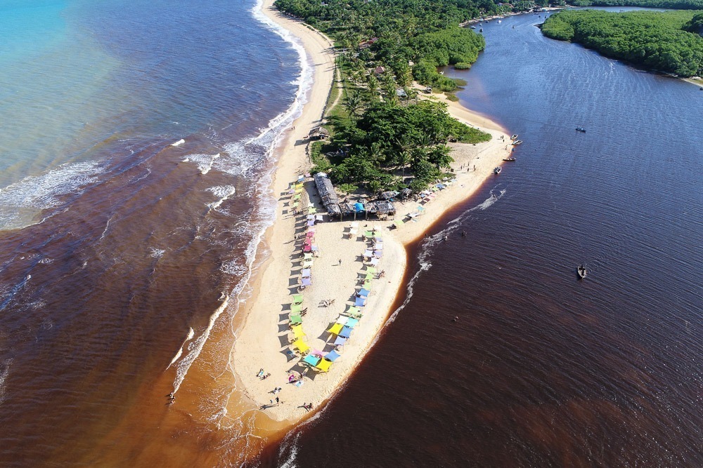 Praia da Barra em Caraíva vista do alto, com barracas coloridas na areia entre rio e mar.