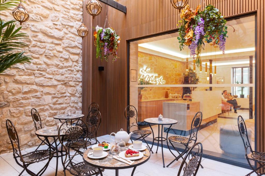 Pátio do Hotel Le Milie Rose, com mesas de ferro, plantas suspensas e área interna vista pela janela