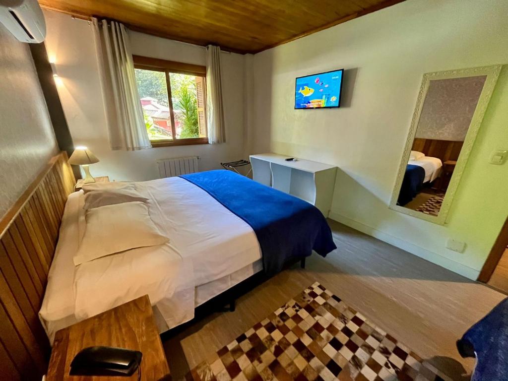 Quarto da Pousada Rosa, em Gramado, com cama de casal, manta azul, televisão e janela voltada para o jardim