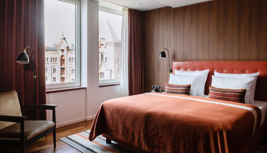 Quarto espaçoso do AMERON Hamburg Hotel Speicherstadt com cama de casal arrumada em tons terrosos e grandes janelas para a cidade.