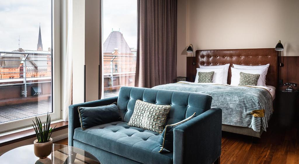 Quarto de hotel elegante e aconchegante com sofá de veludo azul em primeiro plano e cama de casal ao fundo, próximo a uma janela com vista urbana em Hamburgo.