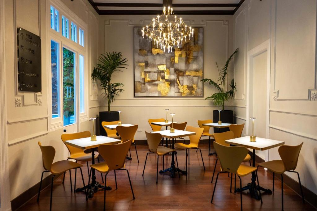 Sala de café da manhã do H Ocoranza com mesas quadradas, cadeiras amarelas, lustre dourado e quadro abstrato dourado na parede.