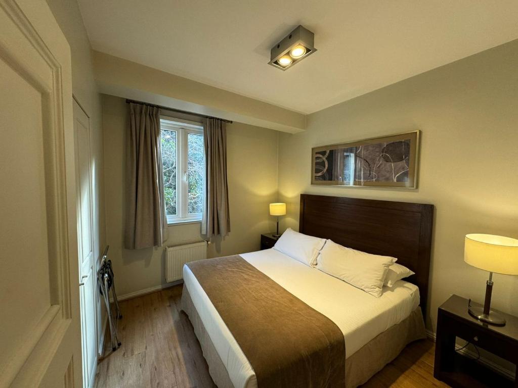 Quarto do Lastarria Aparts com cama de casal, roupa de cama branca e marrom, janela com cortinas e criados de madeira escura.