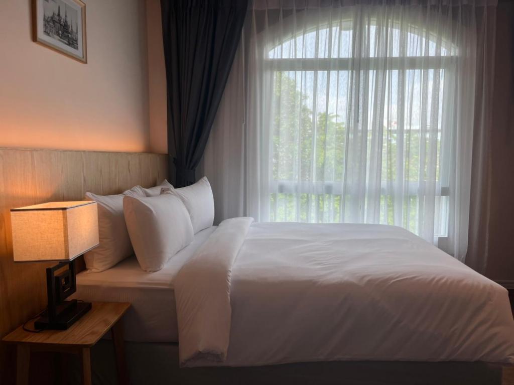 Quarto do AGO HOTEL com cama arrumada com lençóis brancos, uma grande janela ao fundo e abajur sobre mesa de cabeceira de madeira.