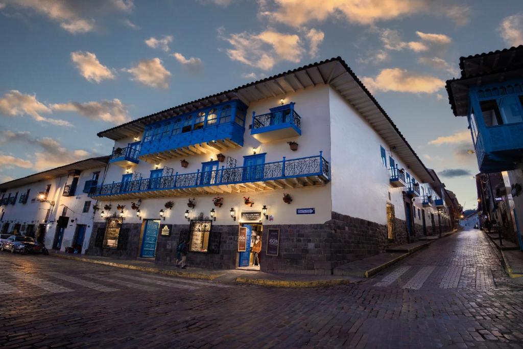 Fachada do Hotel Hacienda Cusco Plaza com paredes brancas, varandas de madeira azul e iluminação nas paredes numa esquina de paralelepípedos.