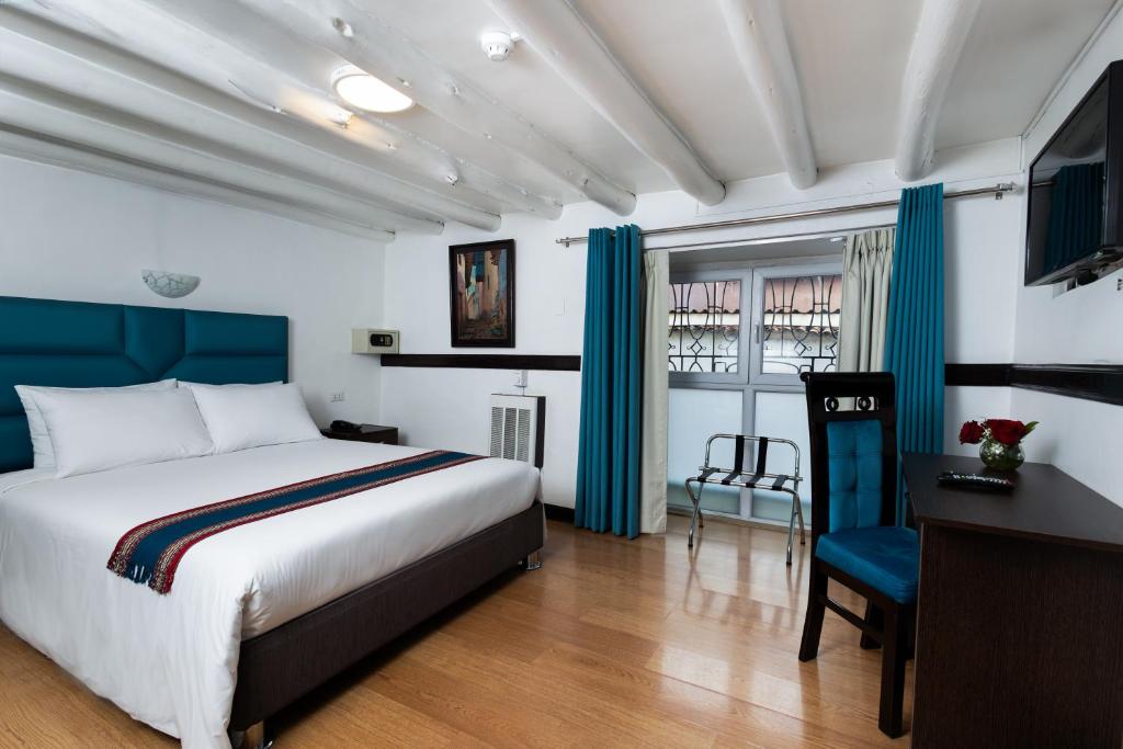 Quarto do Hotel Hacienda Cusco Plaza com cama de casal, cabeceira azul, secretária com cadeira e janela ao fundo.