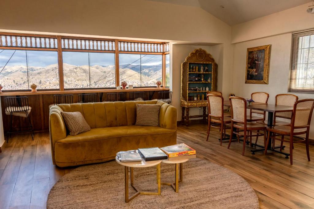 Sala de convívio com sofá de veludo mostarda, mesa de jantar para seis pessoas, mesas de centro e grande fileira de janelas com vista para cidade montanhosa.