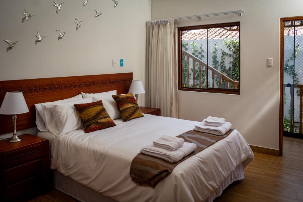 Quarto de casal com cama central, cabeceira de madeira entalhada, toalhas dobradas, abajures em mesinhas de cabeceira e vista para escadaria externa com plantas.