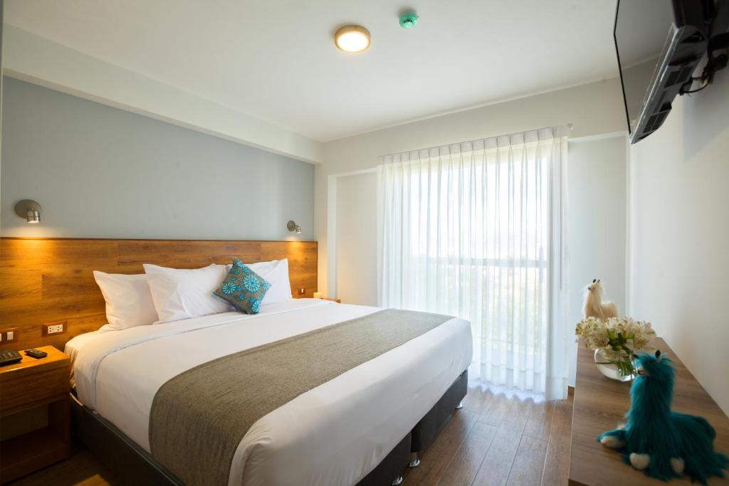 Quarto de hotel moderno com cama de casal grande, cabeceira de madeira, e janela com cortinas brancas.